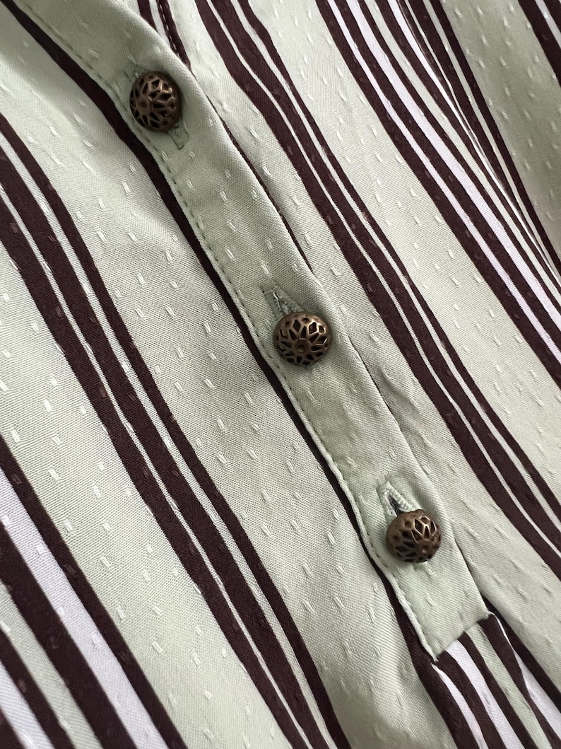 May include: Close-up of a light green and brown striped fabric with decorative metal buttons. The fabric has a subtle textured pattern.