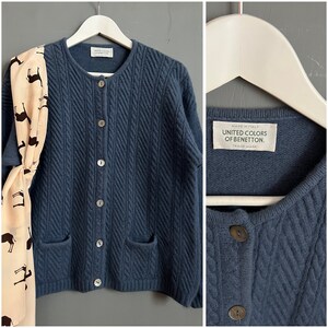 May include: A dark blue cable knit cardigan with a button closure and two pockets. The cardigan has a label that reads "Made in Italy, United Colors of Benetton, Trade Mark."