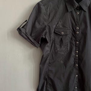 May include: A dark gray button-down shirt with short sleeves, two chest pockets, and a collar. The sleeves are rolled up and secured. The shirt has a floral design on the left chest pocket and a series of buttons down the front.