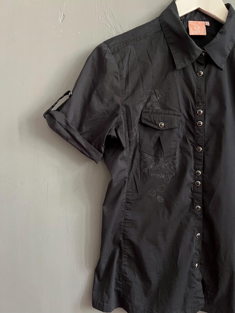 May include: A dark gray button-down shirt with short sleeves, two chest pockets, and a collar. The sleeves are rolled up and secured. The shirt has a floral design on the left chest pocket and a series of buttons down the front.