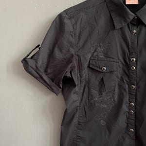 May include: Black short-sleeve button-up shirt with two chest pockets and a collar. The shirt has a floral design on the front and the sleeves are rolled up. The Rip Curl logo is visible on the tag.