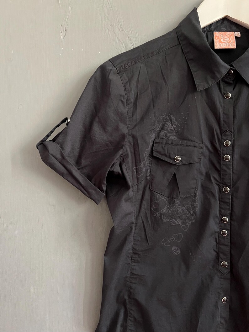 May include: Black short-sleeve button-up shirt with two chest pockets and a collar. The shirt has a floral design on the front and the sleeves are rolled up. The Rip Curl logo is visible on the tag.