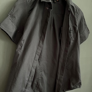 May include: A dark grey, short-sleeved shirt with vertical stripes. The shirt is unbuttoned, revealing the inside. The collar is visible, and the shirt hangs on a white hanger. The brand name "Ben Sherman" is visible on the label.