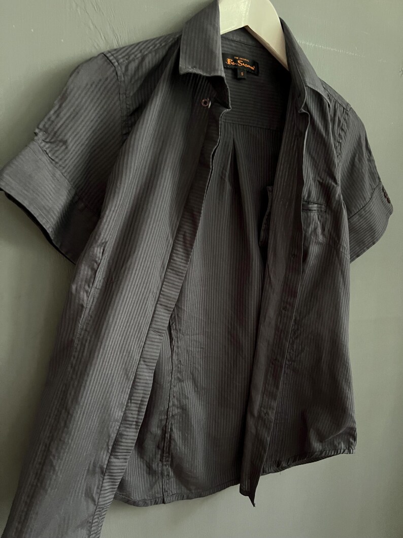 May include: A dark grey, short-sleeved shirt with vertical stripes. The shirt is unbuttoned, revealing the inside. The collar is visible, and the shirt hangs on a white hanger. The brand name "Ben Sherman" is visible on the label.