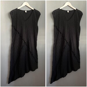 Pode incluir: Um vestido preto sem mangas com decote em V e um acabamento de tecido texturizado e enrugado. A peça apresenta um detalhe de costura diagonal na frente, criando uma bainha assimétrica. É exibido num cabide branco.