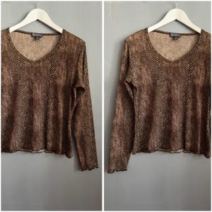 May include: A long-sleeved, brown and black snakeskin-print top with a V-neck. The top is hanging on a white hanger against a gray background. The fabric has a sheer appearance and a ruffled hem.