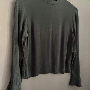 May include: A long-sleeved, dark green blouse with a high neckline and flared sleeves. The blouse is hanging on a white wooden hanger against a gray background. The fabric appears to be a soft, flowing material.