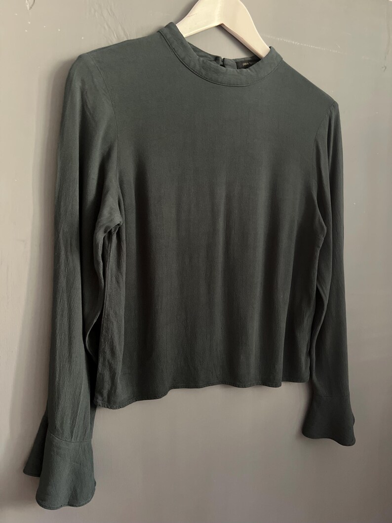 May include: A long-sleeved, dark green blouse with a high neckline and flared sleeves. The blouse is hanging on a white wooden hanger against a gray background. The fabric appears to be a soft, flowing material.