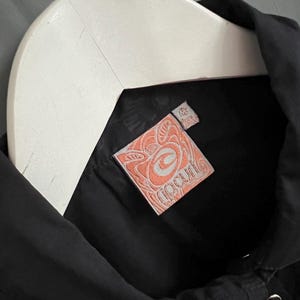 May include: Black button-up shirt with a white hanger. The shirt features a peach-colored tag with the brand name "Ripcurl" in a stylized font. The tag has a decorative design around the brand name.