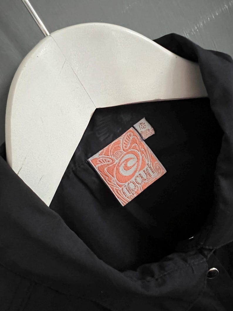 May include: Black button-up shirt with a white hanger. The shirt features a peach-colored tag with the brand name "Ripcurl" in a stylized font. The tag has a decorative design around the brand name.