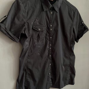 May include: Black short-sleeved button-down shirt with two chest pockets and a collar. The shirt has a subtle floral design on the front and is hanging on a white hanger. The sleeves are rolled up, and the shirt is buttoned up.