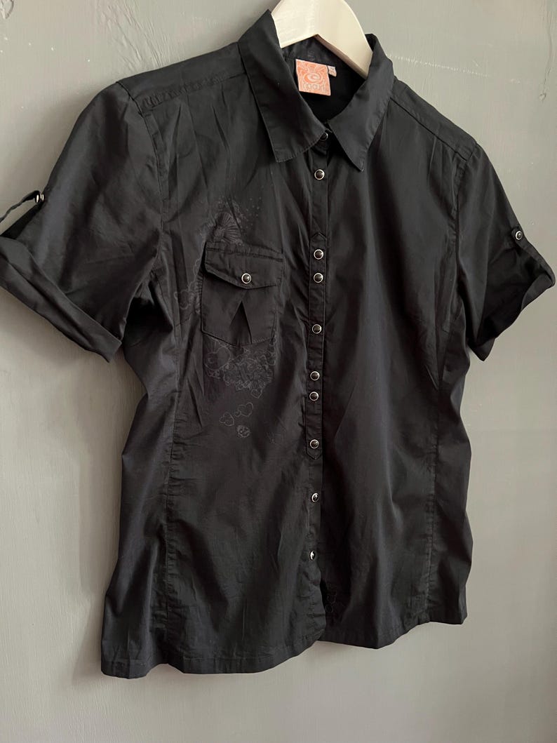 May include: Black short-sleeved button-down shirt with two chest pockets and a collar. The shirt has a subtle floral design on the front and is hanging on a white hanger. The sleeves are rolled up, and the shirt is buttoned up.