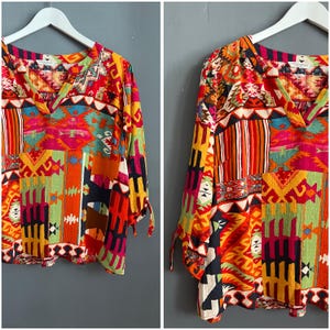 Könnte beinhalten: Ein farbenfrohes Oberteil mit langen Ärmeln, V-Ausschnitt und Bindedetails an den Ärmeln. Das Oberteil hat ein Patchwork-Design mit geometrischen Mustern in Rot-, Orange-, Gelb-, Grün- und Blautönen. Das Kleidungsstück hängt an einem weißen Kleiderbügel.