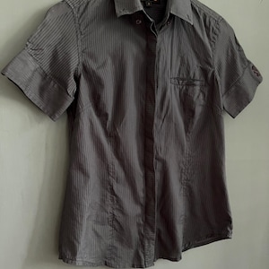 May include: A charcoal grey, short-sleeved button-down shirt with vertical stripes. The shirt features a collared neckline, a chest pocket, and is suspended on a white hanger. The sleeves are cuffed.