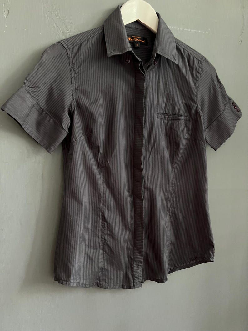 May include: A charcoal grey, short-sleeved button-down shirt with vertical stripes. The shirt features a collared neckline, a chest pocket, and is suspended on a white hanger. The sleeves are cuffed.