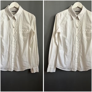 May include: Cream-coloured button-down shirt with long sleeves, a collared neckline, and a chest pocket. The shirt has a subtle dotted pattern and vertical textured stripes down the front. The inside collar features a striped fabric.