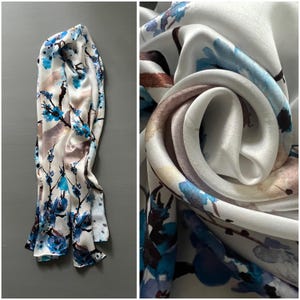 May include: A white silk scarf with a floral pattern in shades of blue, brown, and gray. The scarf is draped and folded, showcasing the soft, flowing fabric. The design features delicate branches and blossoms.