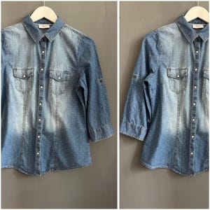 May include: Light blue denim shirt with white polka dots, two chest pockets, and button-down closure. The shirt has three-quarter sleeves and a classic collar. The shirt is hanging on a white hanger.