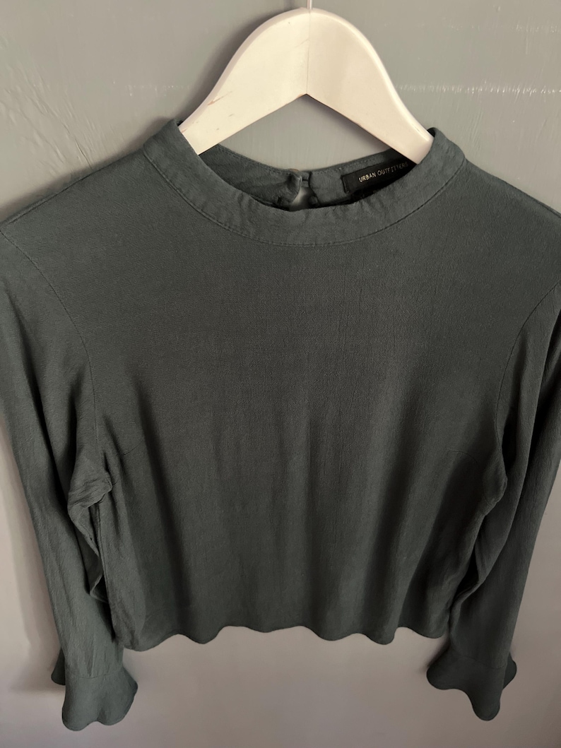 May include: A long-sleeved, dark teal top with a high neckline and flared cuffs. The top is hanging on a white hanger. The tag reads "Urban Outfitters".
