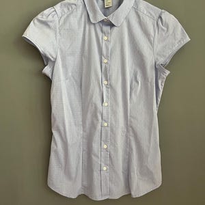 Op de afbeelding: Lichtblauw overhemd met korte mouwen, een ronde kraag en verticale strepen. Het overhemd is dichtgeknoopt en hangt aan een witte hanger. Het overhemd heeft een getailleerde vorm en is gemaakt van een lichte stof.