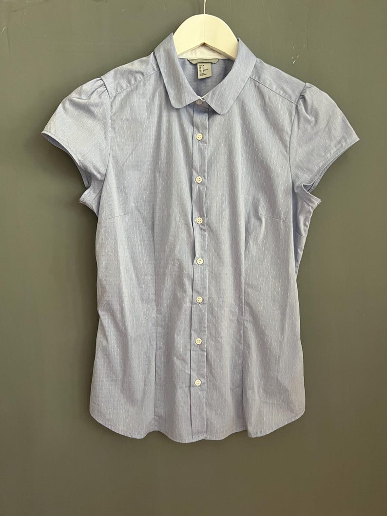 Op de afbeelding: Lichtblauw overhemd met korte mouwen, een ronde kraag en verticale strepen. Het overhemd is dichtgeknoopt en hangt aan een witte hanger. Het overhemd heeft een getailleerde vorm en is gemaakt van een lichte stof.