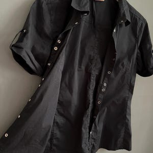May include: Black short-sleeved button-down shirt with a collared neckline. The shirt has silver-colored buttons and roll-up sleeves. The shirt is hanging on a white wooden hanger.