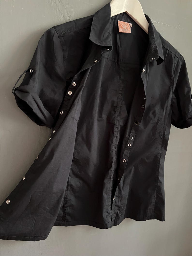 May include: Black short-sleeved button-down shirt with a collared neckline. The shirt has silver-colored buttons and roll-up sleeves. The shirt is hanging on a white wooden hanger.