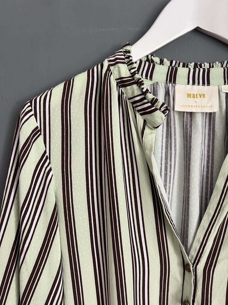 May include: A green and brown striped shirt with a ruffled collar and a button closure. The shirt has a small white tag with the text "Maeve by Anthropologie" on it.