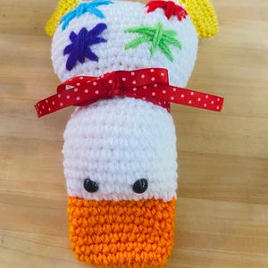 May include: A handmade crochet duck toy. It has a white body, orange bottom, and yellow ears. The duck has black bead eyes and is decorated with colourful embroidered star shapes. A red polka dot ribbon is tied around the neck.