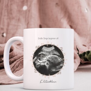 May include: White ceramic mug with a black and white ultrasound image in a floral wreath. The text "Große Dinge beginnen oft Klitzeklein" is printed on the mug.