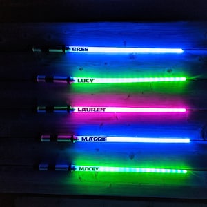Peut inclure: Cinq &eacute;p&eacute;es lumineuses color&eacute;es avec les noms "Bree", "Lucy", "Lauren", "Maggie" et "Mikey" &eacute;crits dessus. Les &eacute;p&eacute;es sont dispos&eacute;es verticalement sur une surface en bois.