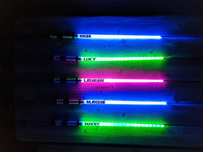 Peut inclure: Cinq &eacute;p&eacute;es lumineuses color&eacute;es avec les noms "Bree", "Lucy", "Lauren", "Maggie" et "Mikey" &eacute;crits dessus. Les &eacute;p&eacute;es sont dispos&eacute;es verticalement sur une surface en bois.