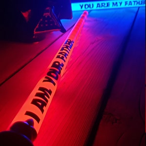 Peut inclure: Un sabre laser rouge avec le texte "I AM YOUR FATHER" dessus, pos&eacute; sur une surface en bois. Un sabre laser bleu avec le texte "YOU ARE MY FATHER" est en arri&egrave;re-plan.