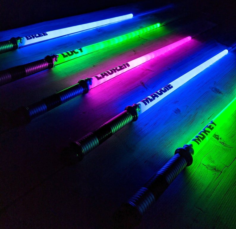 Puede incluir: Cinco espadas de juguete luminosas de colores con nombres escritos en ellas: Bree, Lucy, Lauren, Maddie y Mikey.