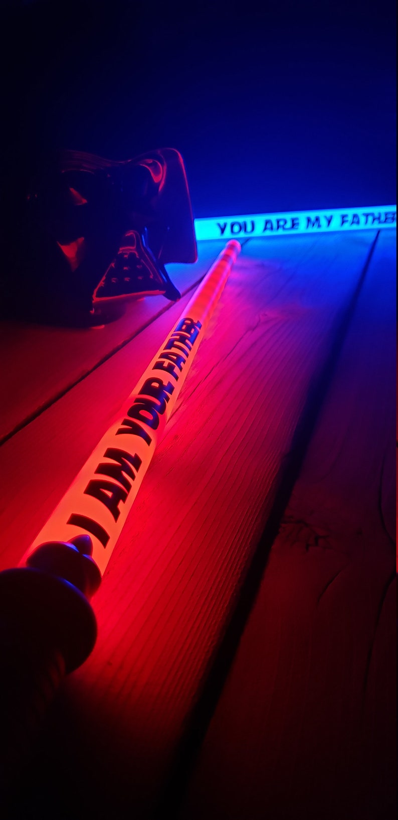 Puede incluir: Un sable de luz rojo con el texto "I AM YOUR FATHER" en &eacute;l, acostado sobre una superficie de madera. Un sable de luz azul con el texto "YOU ARE MY FATHER" est&aacute; en el fondo.