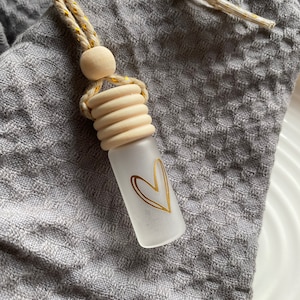Könnte beinhalten: Kleine, mattierte Glasflasche mit goldenem Herzdesign, aufgehängt an einer beige-goldenen Kordel. Die Flasche hat einen Holzverschluss und eine Holzperle. Der Hintergrund ist ein strukturiertes graues Gewebe.