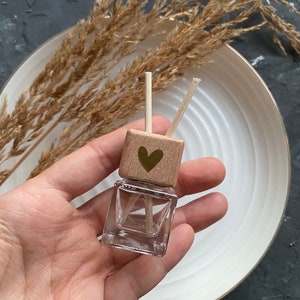 Könnte beinhalten: Kleine, klare Glasflasche mit einem Holzdeckel mit goldenem Herzdesign. Zwei Holzstäbchen ragen aus dem Deckel heraus. Die Flasche wird in einer Hand gehalten, mit einem weißen Teller und getrockneten Pflanzen im Hintergrund.