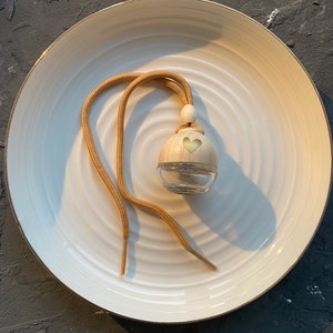 Könnte beinhalten: Eine transparente Glasflasche mit Holzdeckel und herzförmigem Ausschnitt, aufgehängt an einer beigefarbenen Kordel. Die Flasche steht auf einem weißen Teller mit Goldrand. Der Hintergrund ist eine dunkle, strukturierte Oberfläche. Es ist ein Lufterfrischer.