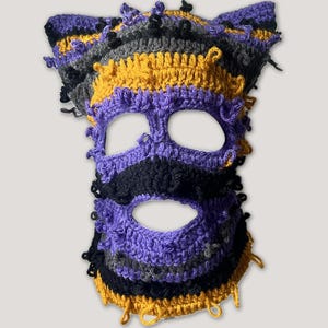 May include: A crochet balaclava featuring a textured, multi-colored design in purple, mustard yellow, black, and gray. The headgear includes two pointed ear-like protrusions at the top and three distinct openings for the eyes and mouth area.