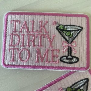 May include: Pink embroidered patch with white stitching. The patch features a martini glass with green olives and the text "Talk Dirty To Me".