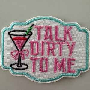 May include: Embroidered patch with a white background, a teal border, and the phrase "TALK DIRTY TO ME" in pink. A pink cocktail with a black stem and a green straw is on the left.
