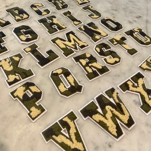 May include: A set of 26 felt letters in a camouflage pattern. The letters are in uppercase and are arranged in alphabetical order. The letters are white with a green and brown camouflage pattern.