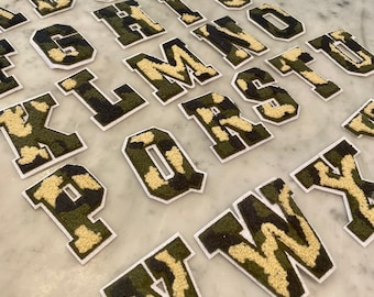 May include: A set of 26 felt letters in a camouflage pattern. The letters are in uppercase and are arranged in alphabetical order. The letters are white with a green and brown camouflage pattern.