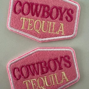 May include: Two pink felt patches with the words "COWBOYS TEQUILA" embroidered in a contrasting color. The patches have a white border and a unique shape, suitable for clothing or accessories.