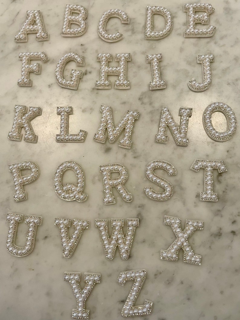 May include: A set of 26 pearl-embellished letter patches in uppercase. The letters are white with a silver outline and are arranged in alphabetical order.