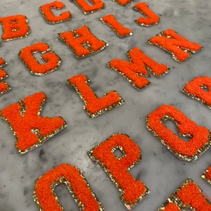 May include: Orange, fuzzy, embroidered letter patches with gold glitter trim. The letters are in a bold, block style. The letters are arranged on a light gray surface. The letters include B, C, D, E, G, H, I, J, K, L, M, N, O, and P.