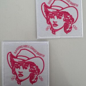 May include: Two white square patches with pink embroidered images of a woman wearing a cowboy hat. The text "Raising Hell Since Birth" is embroidered around the image.