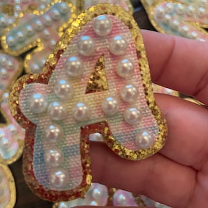 May include: A gold glitter letter A with a rainbow fabric background and white pearl embellishments.