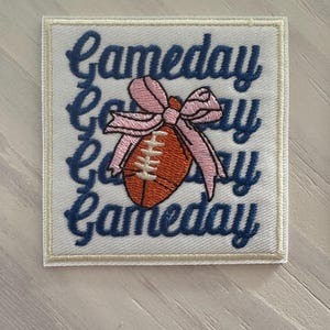 May include: Embroidered patch with the word "Gameday" in navy blue script. A football with white stitching and a pink bow is centered on the patch. The patch has a white background with a cream-colored border.