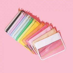 May include: A collection of colorful zippered pouches arranged in a fan shape. The pouches come in a variety of pastel colors, including black, gray, white, light blue, green, yellow, orange, pink, and peach. Each pouch has a gold-colored zipper and a clear front panel.